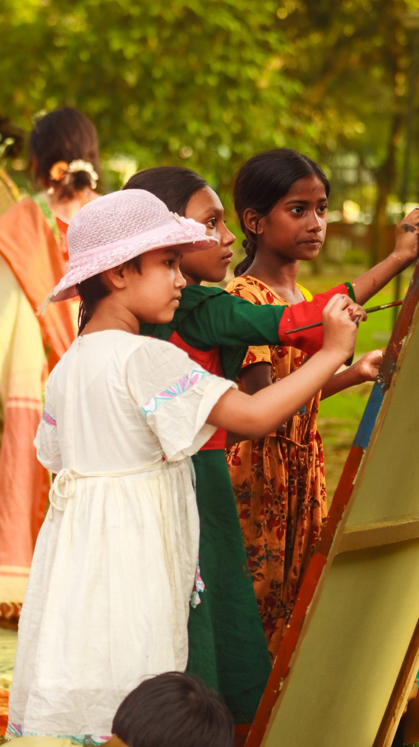 A girl in a white dress and pink hat painting a canvas outdoors alongside other children